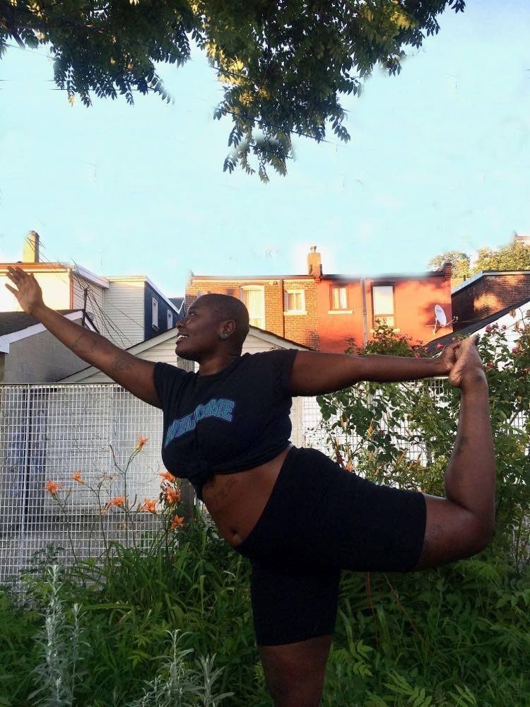 Adwoa, wearing black shorts and a t-shirt, practices Dancer's pose outside among flowers and shrubs.